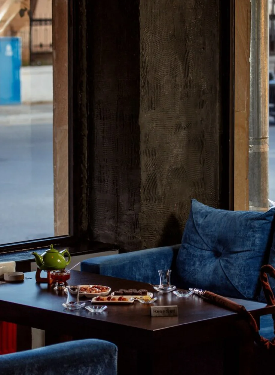 A cozy café corner with a blue velvet couch, a wooden table set with tea, desserts, and a reserved sign by the window.