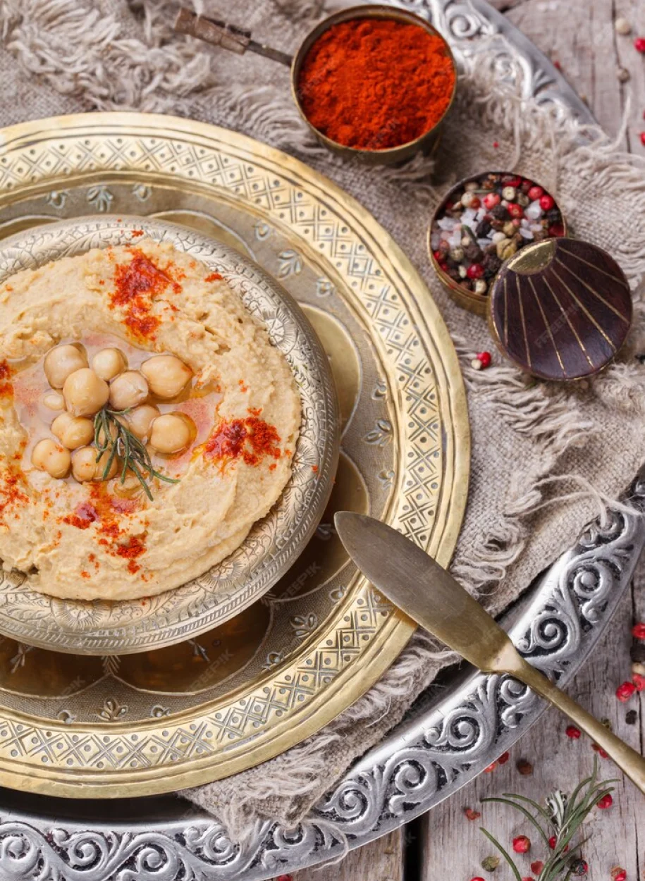 A rich and creamy hummus dish garnished with chickpeas, paprika, and rosemary, served on an ornate metal platter with aromatic spices on the side