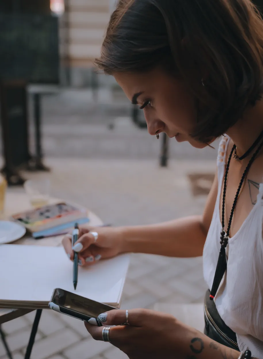 Young woman holding a phone and pen, writing or organizing tasks on paper outdoors.